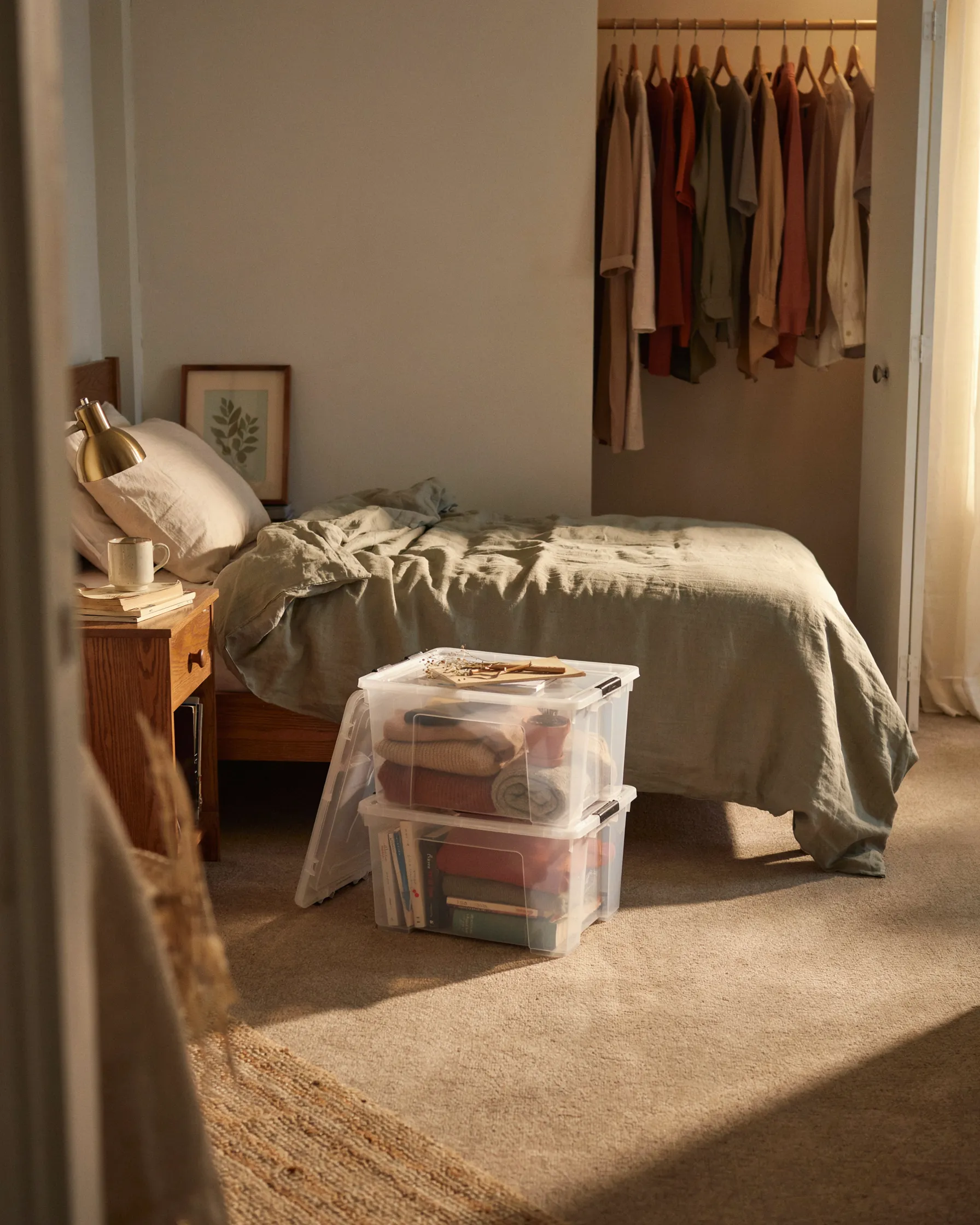 Dorm room corner at golden hour with a made bed, two stacked clear storage bins at the foot of the bed, and hanging clothes visible in an open closet