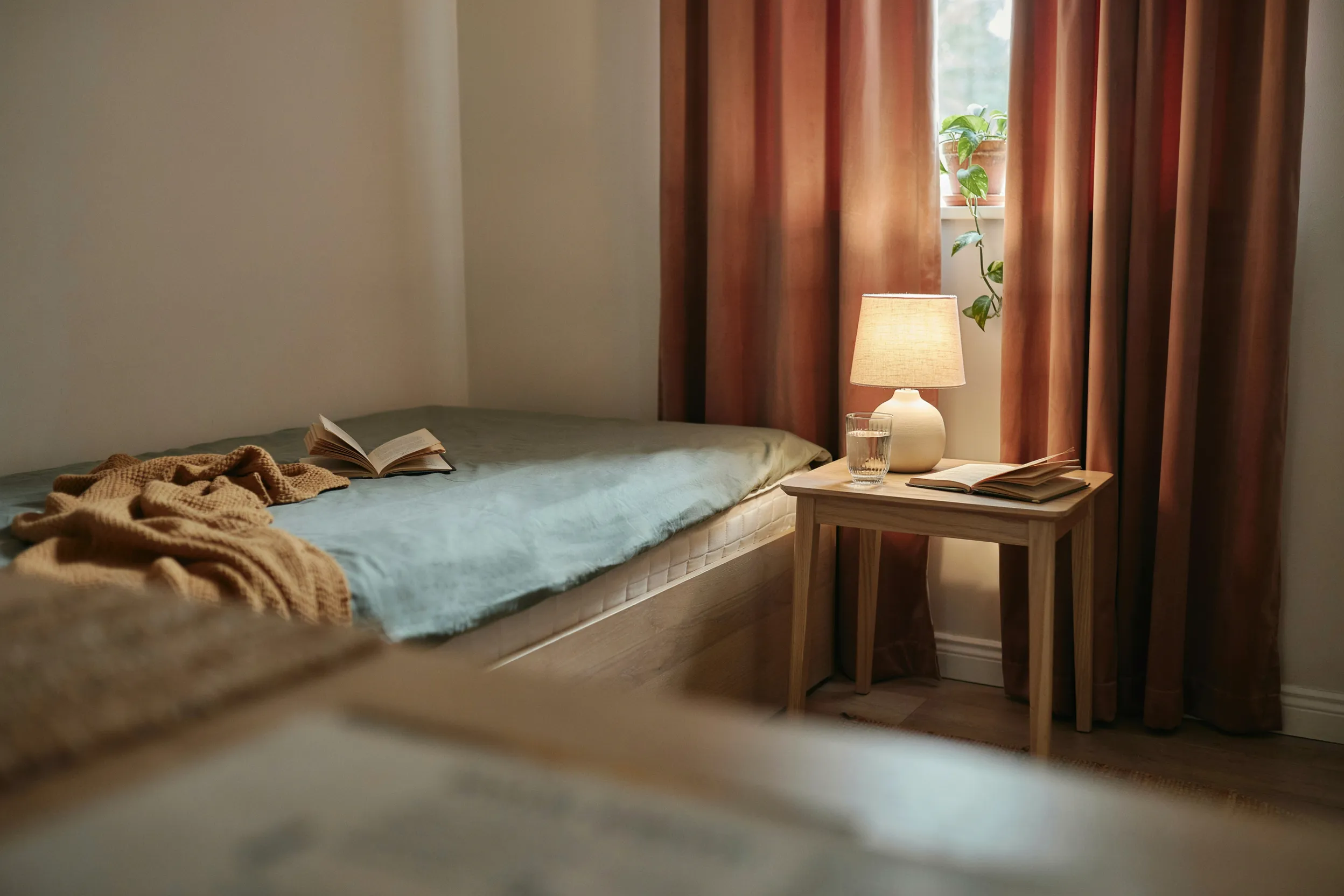 First apartment bedroom corner with a low bed frame, sage bedding, a wooden nightstand with a lamp, and rust-colored curtains